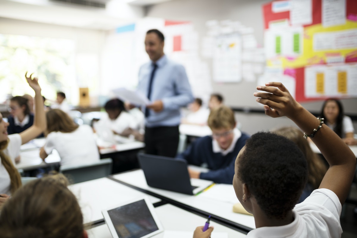 Teacher standing in middle of classroom with young students raising their hands