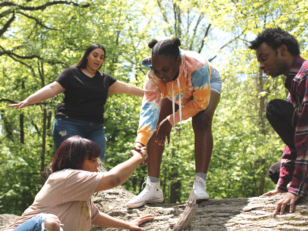 A young woman helps another woman climb up a fallen tree.